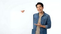 Young Asian man in denim shirt holding blank white sign, smiling and pointing at copy space for advertising or promotional messaging.