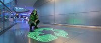 Worker in high-visibility safety vest interacting with glowing green holographic interface on floor in modern futuristic corridor with blue lighting and glass panels.
