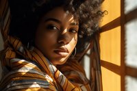 Young African American woman with natural curly hair in striped outfit gazing thoughtfully near window with warm sunlight casting geometric shadows across her face.