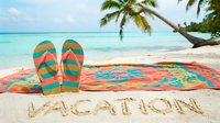 Colorful flip flops and beach towel on tropical sand with vacation written, palm trees and turquoise ocean in background on sunny day.