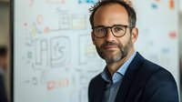 Middle-aged Caucasian businessman with glasses and beard wearing navy suit jacket smiles confidently against blurred office background with strategy diagrams and notes.