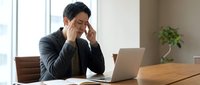 Stressed Asian businessman holding head in hands while working on laptop at office desk, experiencing work pressure and mental fatigue in modern workplace.