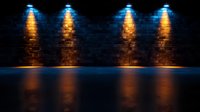 Dramatic dark brick wall illuminated by four orange spotlights casting reflections on glossy floor surface, creating moody atmospheric stage or gallery space.