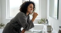 Young African American woman in gray robe brushing teeth in modern bathroom with natural light from window. Morning dental hygiene routine and self-care concept.