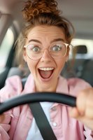 Young Caucasian woman with glasses showing excited expression while driving car, wearing pink jacket. Close up portrait capturing genuine joyful emotion.