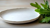 Empty ceramic plate with rustic beige rim on wooden table surface, fresh green arugula leaves beside, minimalist food photography setup for restaurant menu.