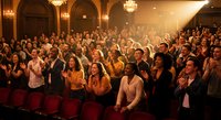 Large diverse audience applauding in elegant theater with ornate architecture and warm lighting during live performance or presentation event.