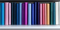 Row of colorful hardcover books arranged on white shelf, spines showing gradient from blue and purple to pink and orange tones, creating minimalist library display.