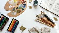 Artist workspace flat lay with wooden palette, colorful paints, brushes, pencils, sketchbook and autumn leaves on white background for creative projects.