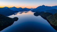 Aerial view of tranquil mountain lake at sunset with deep blue silhouettes of peaks reflecting in calm waters, surrounded by forested peninsulas and islands.