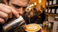 Skilled male barista pouring steamed milk into a latte art cup at a busy coffee shop, showcasing craft and expertise in specialty coffee.