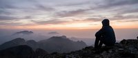 Silhouetted figure sitting on rocky mountain peak at sunset, gazing at distant misty peaks and dramatic cloudy sky during golden hour. Suitable for motivational, travel, and lifestyle content.