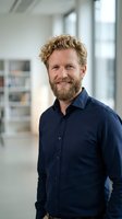 Confident middle-aged Caucasian businessman with curly hair and beard smiling in modern office environment wearing navy blue shirt for corporate headshot.