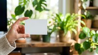 Hand holding blank white card in bright room with potted green plants and natural window light, suitable for business cards, invitations, and promotional designs.
