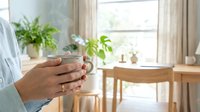 Woman holding glass mug in bright modern kitchen with plants on windowsill. Morning coffee break, healthy lifestyle, home comfort concept.