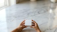 Hands holding blank white box on marble floor with natural light from window, suitable for product packaging mockups and branding presentations.