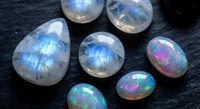 Collection of natural rainbow moonstone and opal gemstones displaying iridescent blue and multicolor flashes on dark slate surface for jewelry making.