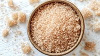 Brown sugar crystals in wooden bowl on white wooden table with scattered sugar cubes, top view macro shot showing grainy texture and natural sweetener.