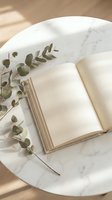 Open book with eucalyptus branches on marble table in natural sunlight creating peaceful reading atmosphere for relaxation and mindfulness.