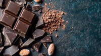 Dark chocolate pieces and cocoa powder scattered on textured blue background with roasted cacao beans, overhead view. Food photography for confectionery marketing.