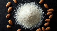 White flour scattered in circle shape with raw almonds arranged around on black stone background, overhead view for food photography and baking concepts.