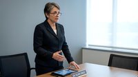 Professional businesswoman in dark suit and glasses standing at conference table in modern office with tablet and documents for corporate meetings.