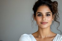 Young woman with natural makeup and curly dark hair wearing white against gray background smiling confidently at camera, radiating warmth and authenticity.