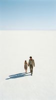 Adult man and woman holding hands walking across vast white salt flat desert under clear blue sky, casting long shadows, suitable for travel, adventure, relationships, and lifestyle editorial use.
