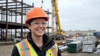 Young Asian female construction worker smiling at building site wearing orange hard hat and safety vest with crane and steel framework in background.
