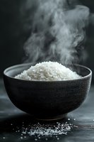 Steaming white rice in black ceramic bowl against dark background, showcasing rising vapor and scattered grains on wooden surface.