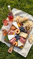 Overhead view of a wooden charcuterie board arranged with cured meats, cheeses, fresh fruits, olives, bread, and rosemary on grass with rose wine for outdoor entertaining.