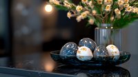 Elegant Easter eggs in navy and gold on dark plate with spring flowers in background create luxurious holiday atmosphere. Soft evening lighting adds warmth.