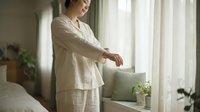 Middle-aged woman with light skin tone wearing beige linen pajamas standing by window with sheer curtains in bright morning light, relaxed home lifestyle scene for wellness and sleep content.