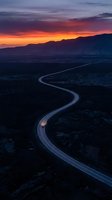Winding highway with vehicle headlights cutting through dark landscape at sunset, dramatic orange and purple sky above mountains, aerial view for travel and transportation concepts.