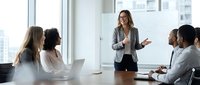 Professional businesswoman presenting to diverse team in modern conference room with city view windows during corporate meeting.