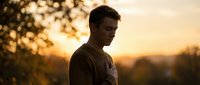 Young adult male with light skin tone and dark hair wearing casual clothing, standing outdoors during golden hour sunset with blurred bokeh background, conveying contemplation and solitude.