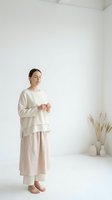 Serene woman with dark hair and East Asian ethnicity wearing layered cream and beige linen clothing standing barefoot in minimalist white interior space with dried pampas grass decor.