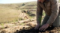 Farmer in brown jacket and gloves working soil in agricultural field with rolling landscape background, suitable for farming, agriculture, and rural lifestyle content.