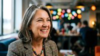 Mature woman with long gray hair and checkered blazer smiling in modern office environment with blurred colleagues and warm lighting in background.