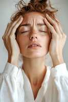 Young woman with freckles and closed eyes touching temples, wearing white shirt, expressing headache, stress or anxiety. Soft natural lighting creates mood.