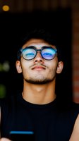 Young Asian man wearing blue light blocking glasses with confident expression against dark background. Portrait captures modern professional lifestyle.