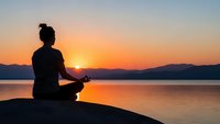 Woman silhouette meditating in lotus pose at sunrise by calm lake with mountains. Peaceful morning yoga practice for mindfulness and spiritual wellness.