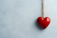 Red ceramic heart ornament hanging on rope against light blue textured wall background for romantic decoration and love concept design projects.