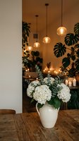 Modern dining room interior with white hydrangea flowers in ceramic vase on wooden table, pendant lights and tropical plants creating cozy atmosphere.