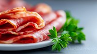 Thinly sliced prosciutto arranged on white plate with fresh parsley garnish, macro shot with selective focus showing meat texture and marbling.