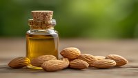 Golden almond oil in glass bottle with cork stopper surrounded by raw almonds on wooden surface with natural green background for skincare.