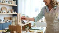 Middle-aged woman with light skin tone and blonde hair, wearing a beige apron over a green shirt, arranging wooden home decor items and potted plants on a shelf in a bright, modern kitchen with blue cabinetry.
