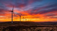Wind turbines generating renewable energy against dramatic sunset sky with vibrant orange and purple clouds over rolling landscape.