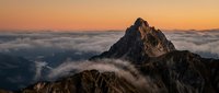 Dramatic mountain peak rising above clouds during golden hour sunset with warm orange sky and misty valleys below creating breathtaking alpine landscape scenery.
