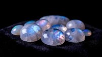 Iridescent rainbow moonstone gems with holographic flash on dark textured fabric surface for jewelry making and gemstone collection displays.